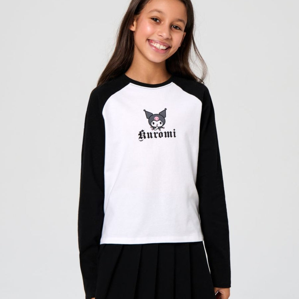 A young girl smiling and wearing a black and white long-sleeve raglan shirt with a cartoon character graphic and the word 'Anomi' on the chest, paired with a black pleated skirt, against a light gray studio background.