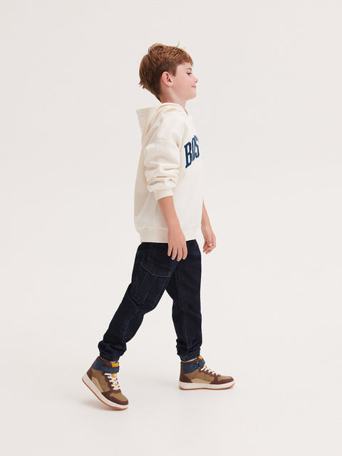 Child wearing a cream hoodie with text and dark jeans on a white background