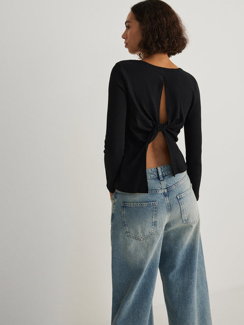 Person wearing a black long-sleeve top with a knot detail at the back and light blue jeans on a plain background