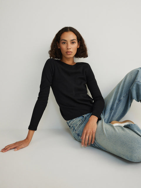 Woman wearing a black sweater and light blue jeans sitting on a white floor.