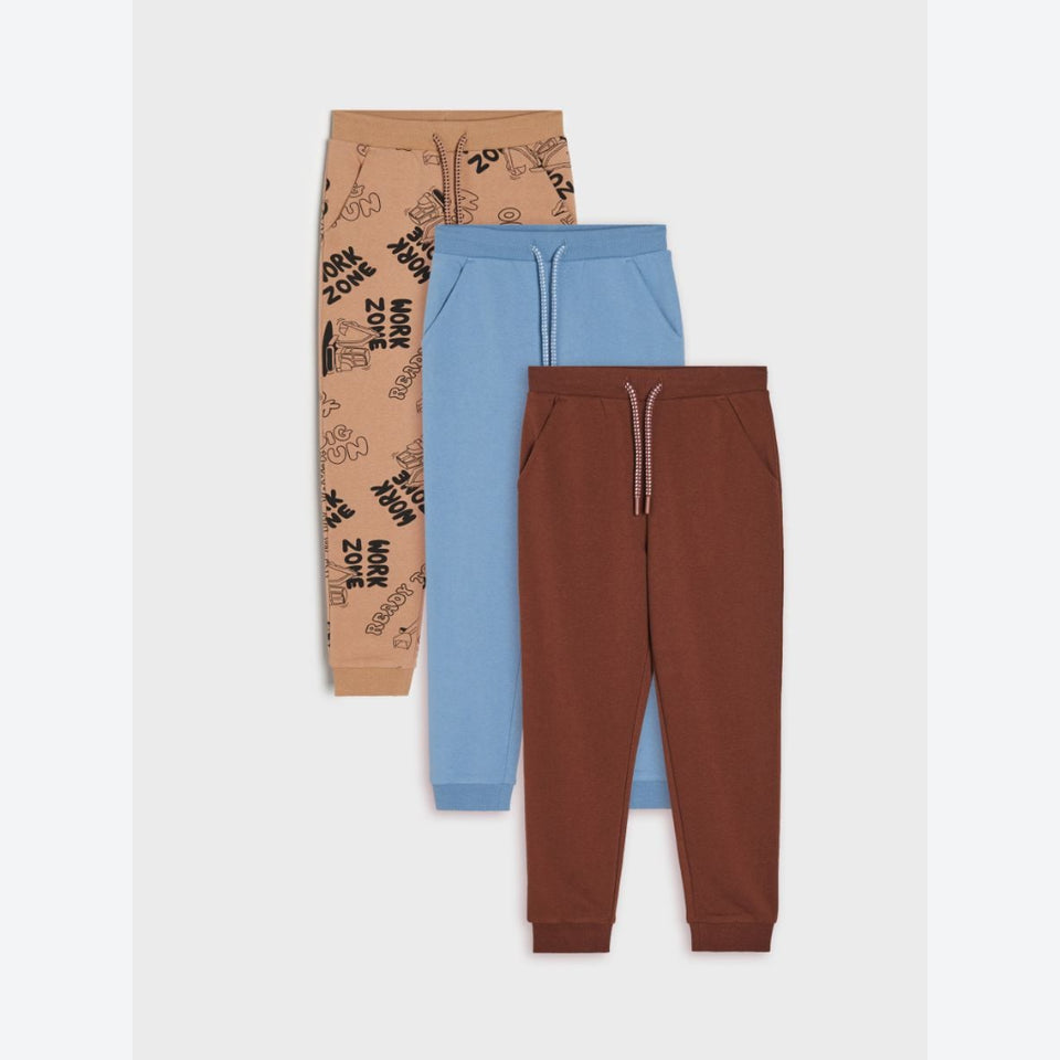 Three pairs of children's jogger pants stacked vertically on a light gray background: tan with black graphic text print, light blue, and rust brown.