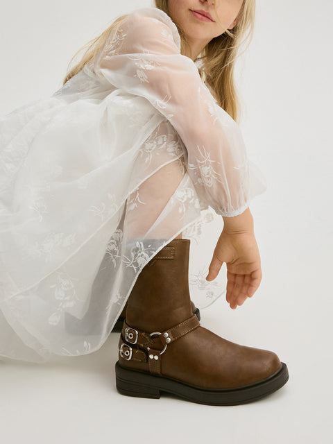 Child wearing a white dress with sheer sleeves and brown boots on a white background