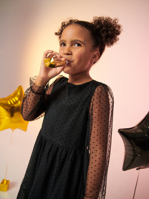 Child in a black dress drinking from a small bottle with balloons in the background