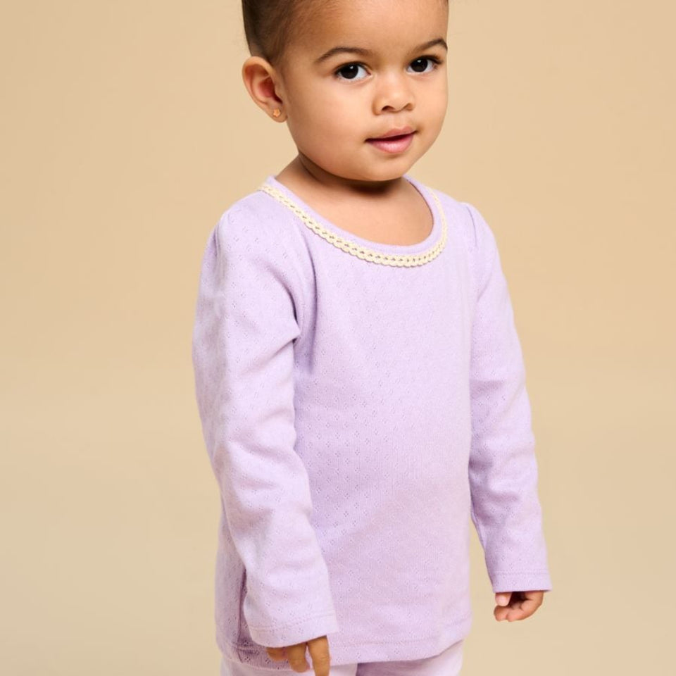 A toddler girl with her hair in a high bun is wearing a light lavender long-sleeve top with a decorative pearl collar trim, paired with matching lavender pants, set against a solid beige background.