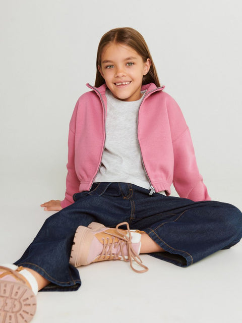 Young girl wearing a pink jacket, white shirt, blue jeans, and pink shoes on a light gray background