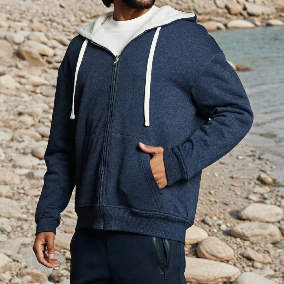 A man wearing a navy blue zip-up hoodie with a white hood lining and white drawstrings, standing on a rocky beach next to the water.