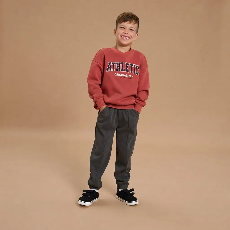 A young boy smiling and posing in a studio setting, wearing a rust-red crewneck sweatshirt with 'ATHLETIC ORIGINAL CITY' printed on the chest, dark gray jogger pants, and black sneakers with white soles, against a solid tan background.