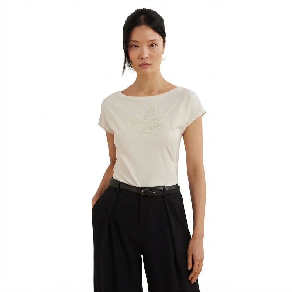 A woman modeling a short-sleeve cream-colored top with a subtle tonal floral embroidery on the chest, paired with black wide-leg trousers and a black belt, against a solid white background.