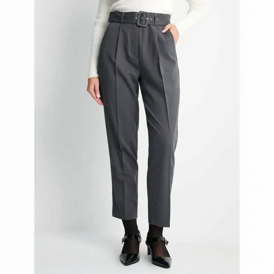 A model wearing high-waisted dark gray pleated trousers with a matching dark gray belt, styled with a white long-sleeve top, black socks, and black patent Mary Jane shoes against a light gray studio background.