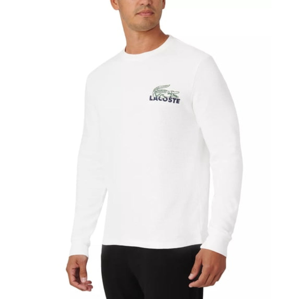 A man wearing a white long-sleeve crewneck shirt with a green and navy Lacoste crocodile logo on the left chest, paired with black pants, against a plain white background.