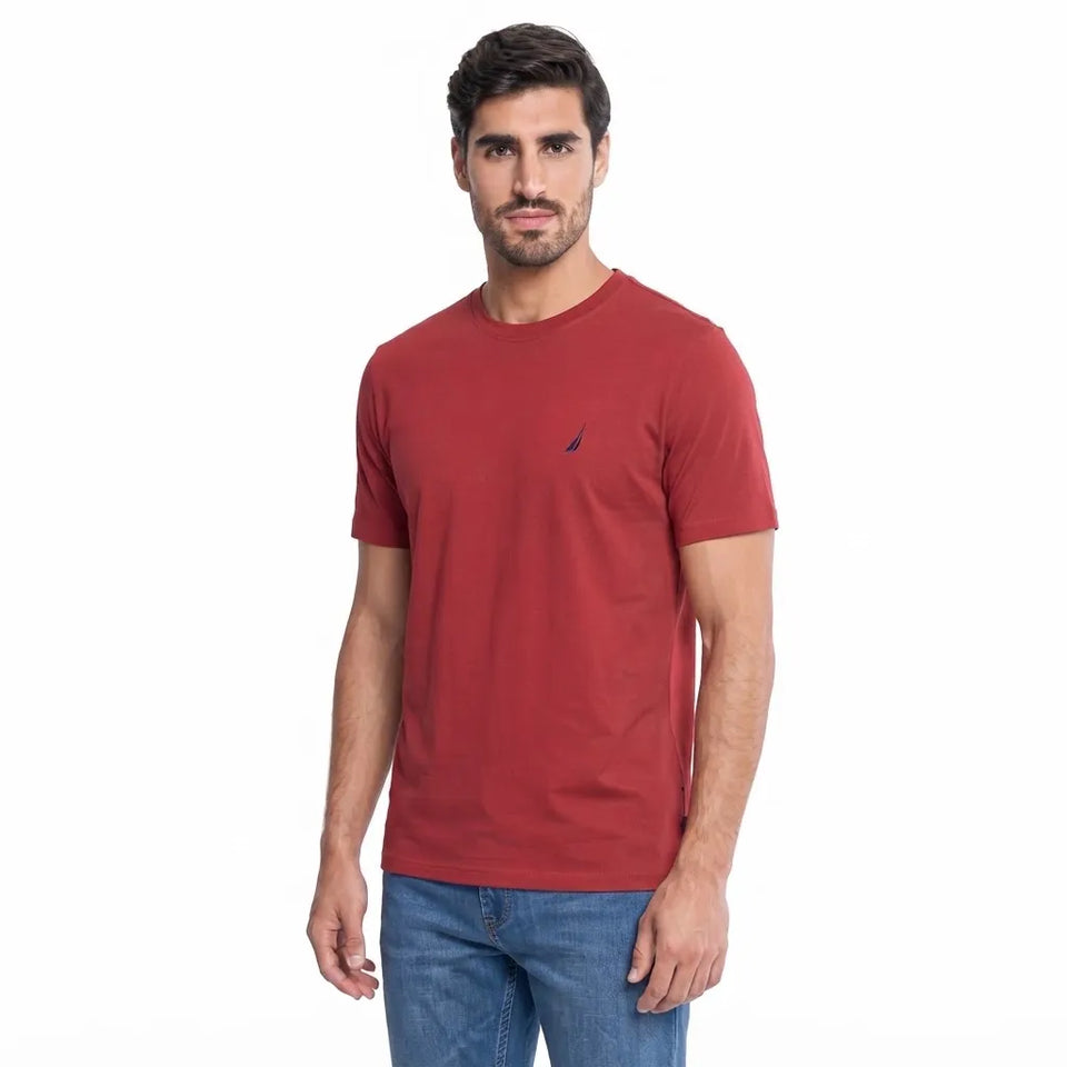 A man wearing a red short-sleeve crewneck t-shirt with a small navy blue sail logo on the left chest, paired with blue jeans, against a plain white background.