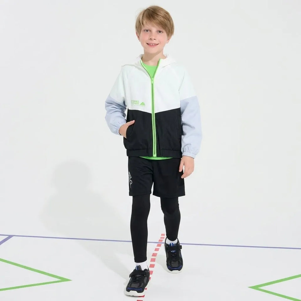 A young boy modeling a color-blocked zip-up jacket, black shorts, and black leggings with athletic shoes, standing on a white studio floor with colored lines.