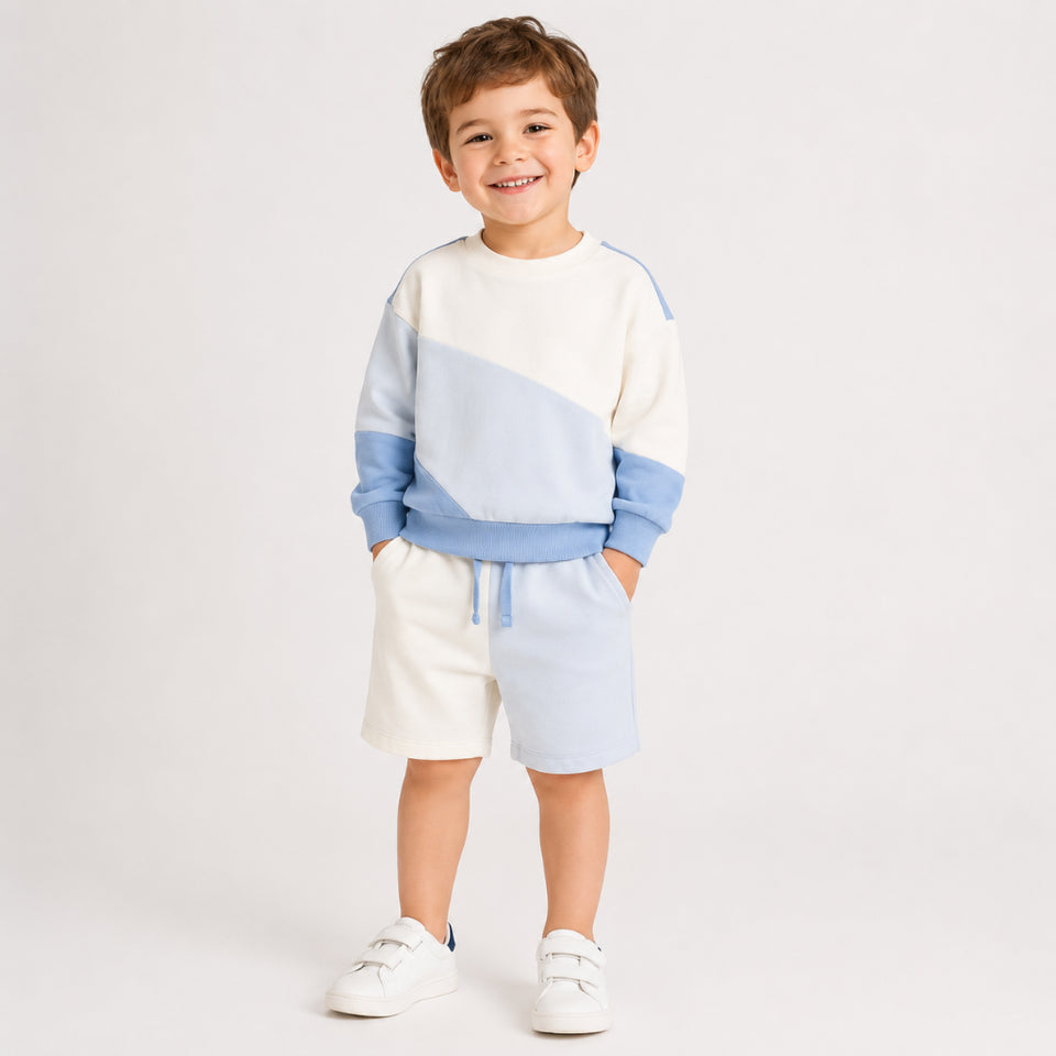 A young boy smiling and wearing a matching color-blocked sweatshirt and shorts set in cream, light blue, and medium blue, paired with white sneakers, against a plain light gray studio background.