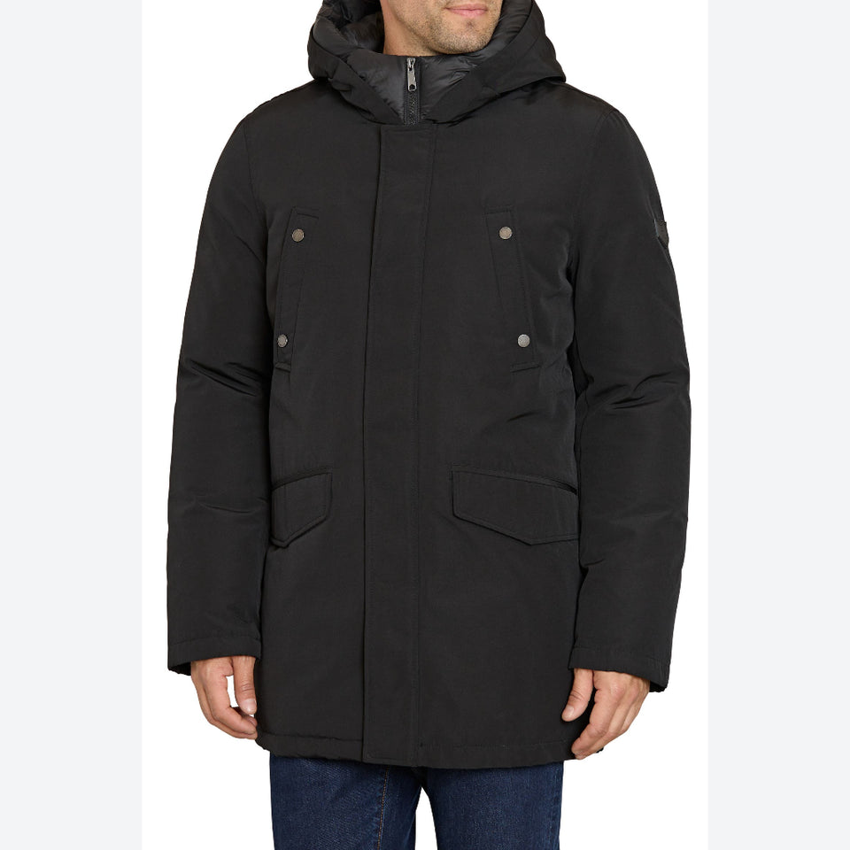 A man wearing a black hooded winter parka with a front zipper, four buttoned flap pockets, and a high collar, paired with blue jeans against a plain white background.