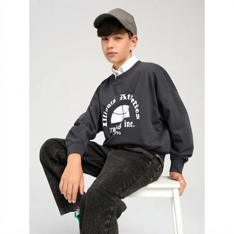 A boy wearing a dark gray crewneck sweatshirt with a white circular graphic that reads 'Xavier Athletics Inc. 1996', paired with black jeans, a white collared shirt, and a gray baseball cap, sitting on a white stool against a light gray background.