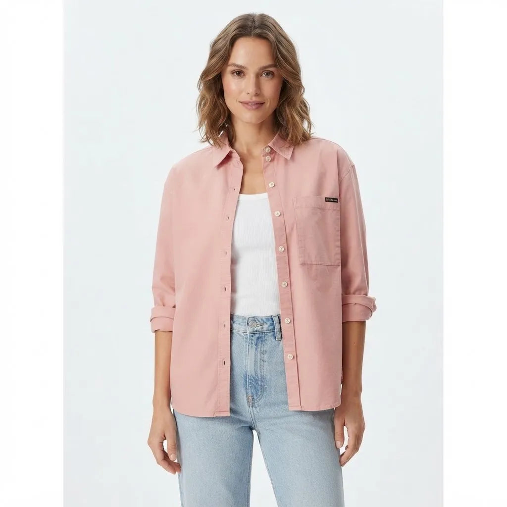 A woman modeling a light pink button-up shirt with rolled-up sleeves, worn open over a white tank top and light-wash blue jeans, against a plain light gray studio background.