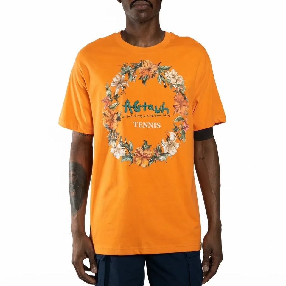 A man wearing a bright orange short-sleeve t-shirt with a circular floral wreath graphic on the chest featuring the words 'Agath Tennis'.