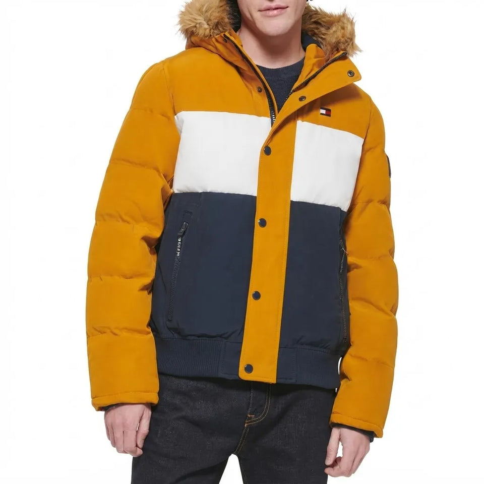 A model wearing a Tommy Hilfiger colorblock puffer jacket with a mustard yellow body, a white chest stripe, a navy blue lower section, and a brown faux-fur hood trim.