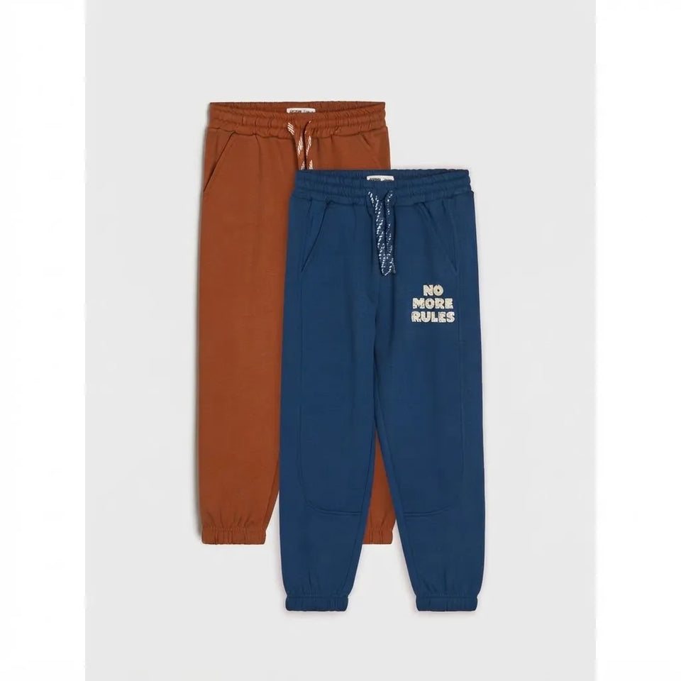 Two pairs of kids' jogger sweatpants, one in rust orange and one in navy blue, displayed flat against a light gray background. The navy pair features white text reading 'NO MORE RULES' on the left thigh.