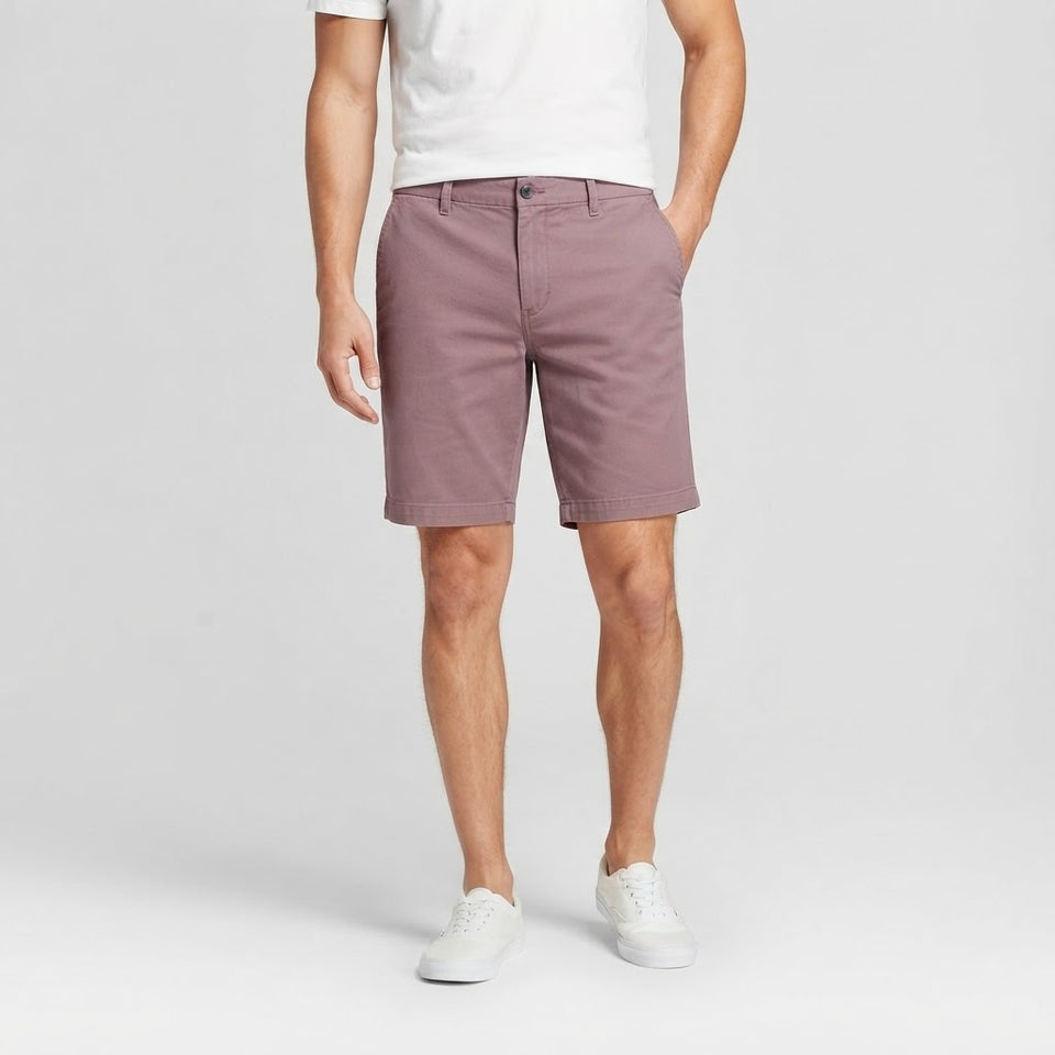 A man wearing dusty rose chino shorts, a white t-shirt, and white sneakers, standing against a light gray studio background.