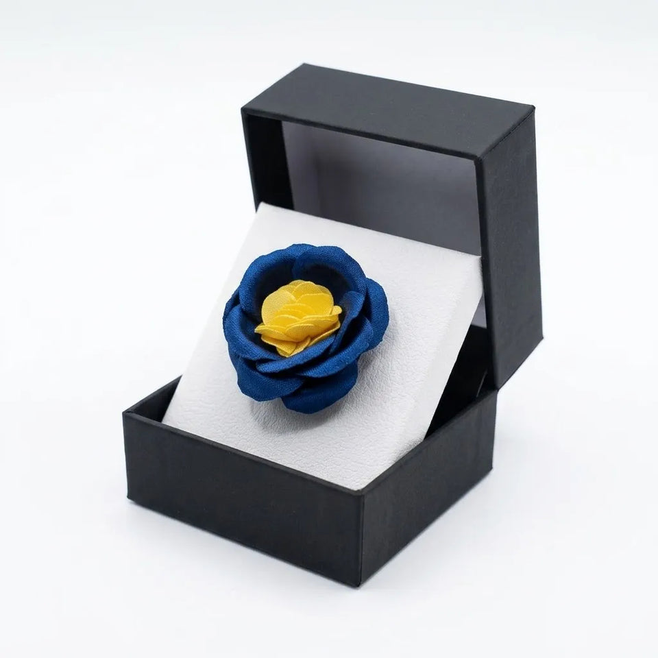 A blue and yellow fabric flower brooch displayed inside an open black presentation box with a white insert, set against a plain white background.