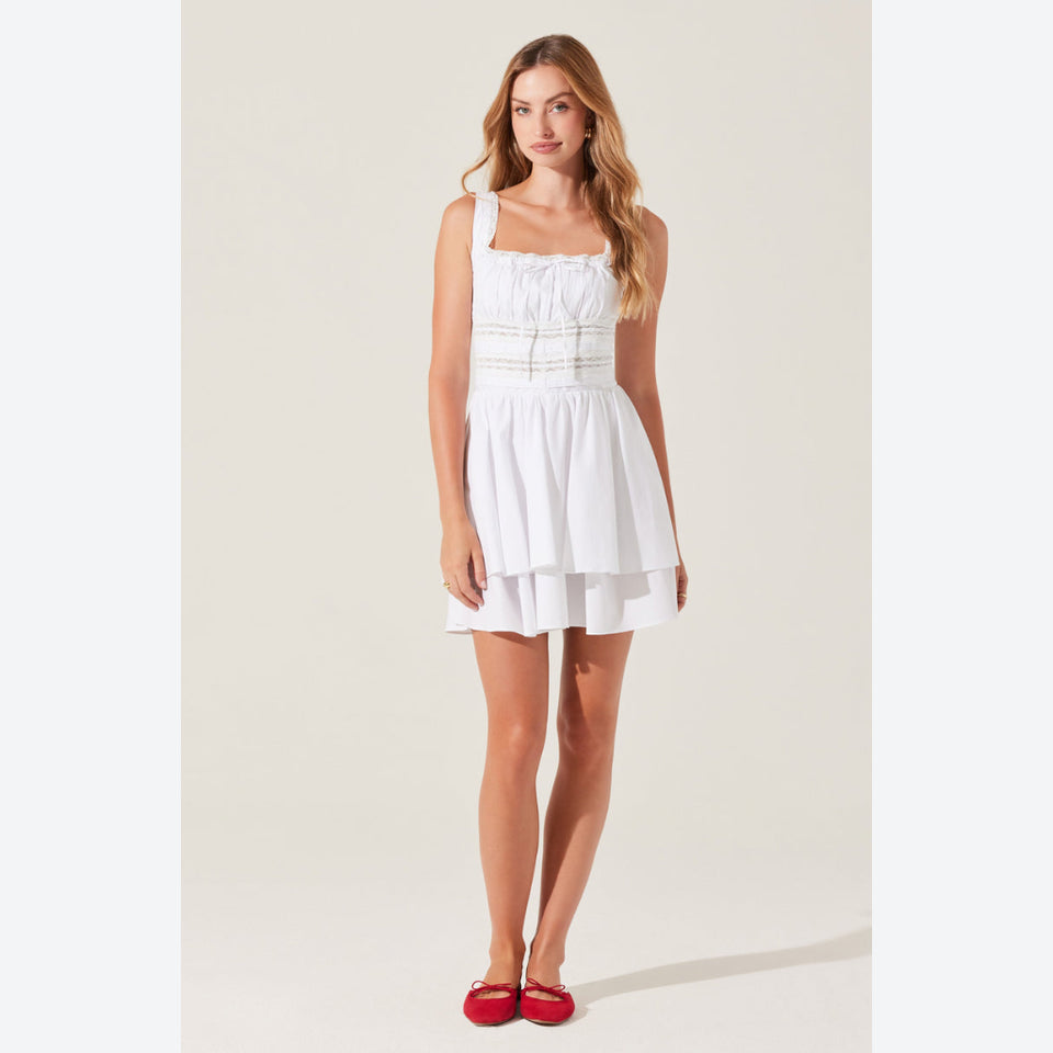 A woman modeling a white sleeveless tiered mini dress with thin straps and horizontal gathered detailing at the bodice, paired with red flats, against a plain light beige studio background.