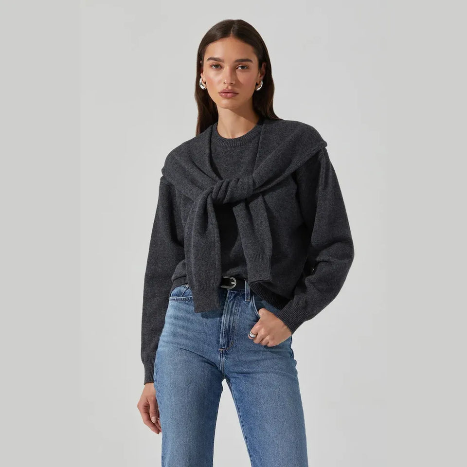 A woman modeling a dark gray knit sweater with a draped, tie-front design, paired with blue jeans, against a solid light gray studio background.