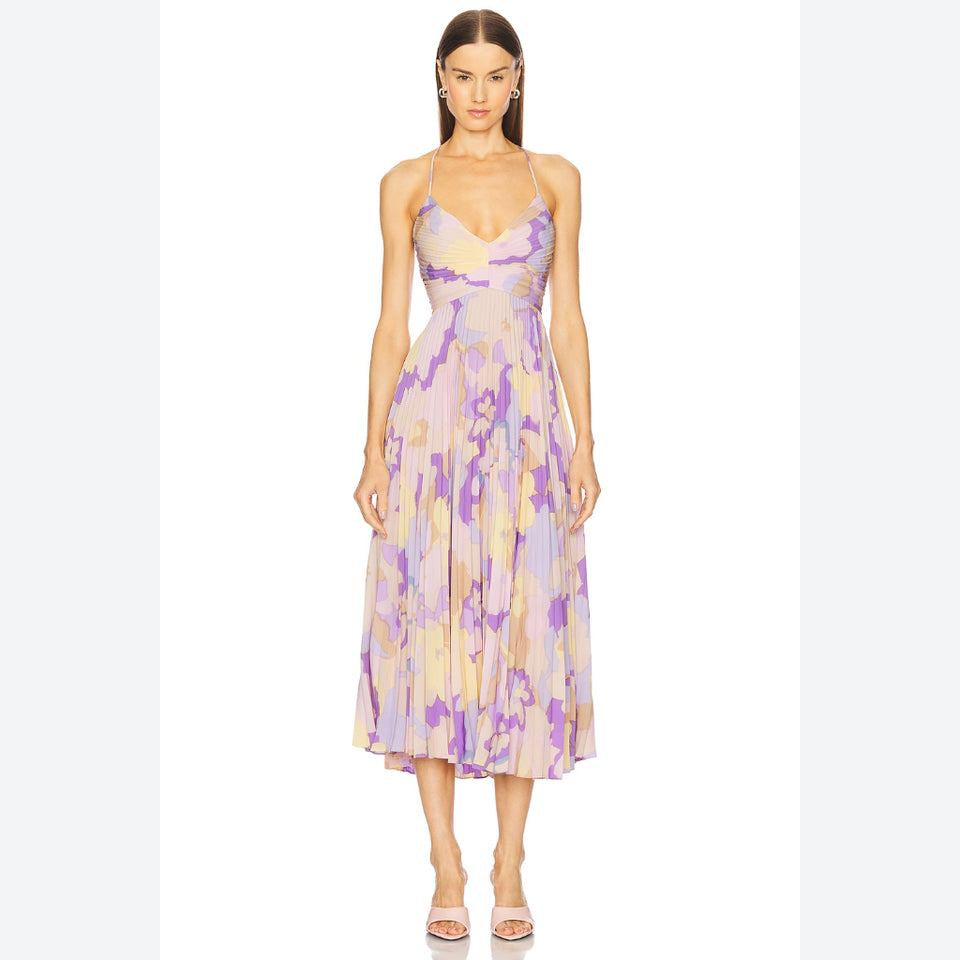A woman modeling a sleeveless midi dress with thin straps and a purple, yellow, and beige abstract watercolor print, standing against a plain white background.