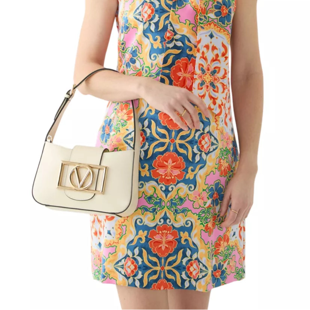 A model wearing a colorful halter mini dress with a vibrant floral and ornamental print, holding a cream-colored shoulder bag with a gold-tone clasp.