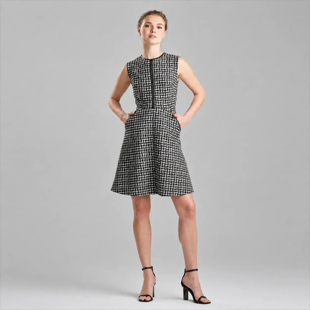 A woman modeling a sleeveless black and white micro-check patterned dress with a V-neckline and front zipper, paired with black high-heeled sandals, against a solid gray studio background.
