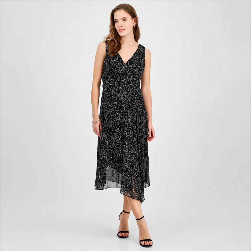 A woman modeling a sleeveless black midi dress with a white abstract floral pattern, featuring a V-neckline and a sheer overlay at the hem, set against a light gray studio background.