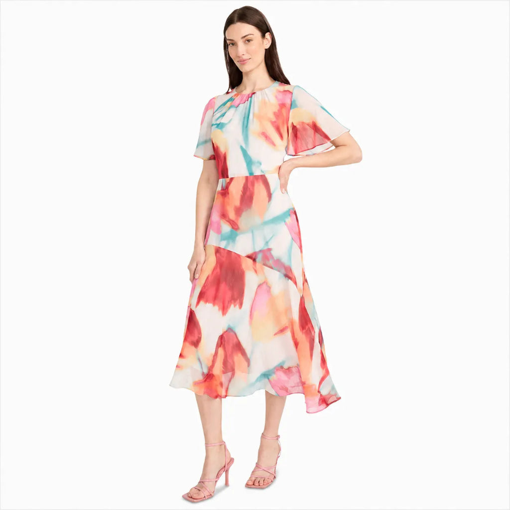 A woman modeling a short-sleeve midi dress with a vibrant watercolor floral print in red, orange, pink, and teal on a white base, paired with light pink high-heeled sandals against a plain white background.