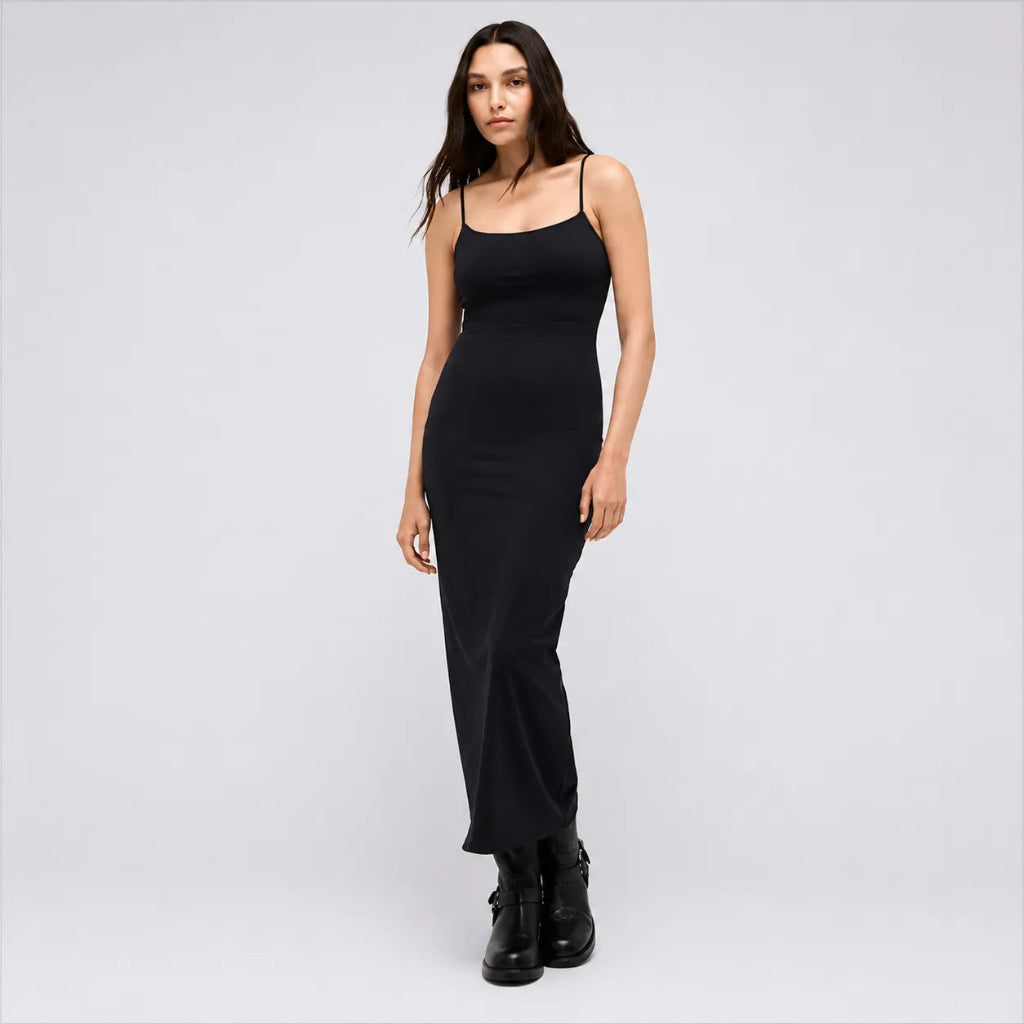 A woman modeling a black spaghetti-strap midi dress with a fitted silhouette, paired with black ankle boots, against a light gray studio background.