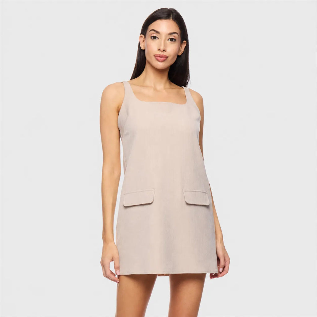A woman modeling a sleeveless beige mini dress with a square neckline and two front flap pockets, set against a solid light gray background.