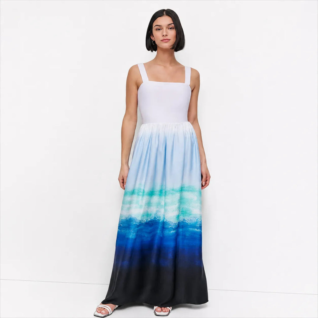 A woman modeling a sleeveless maxi dress with a white bodice and a flowing skirt that gradients from light blue and teal to deep blue and black, set against a plain white studio background.