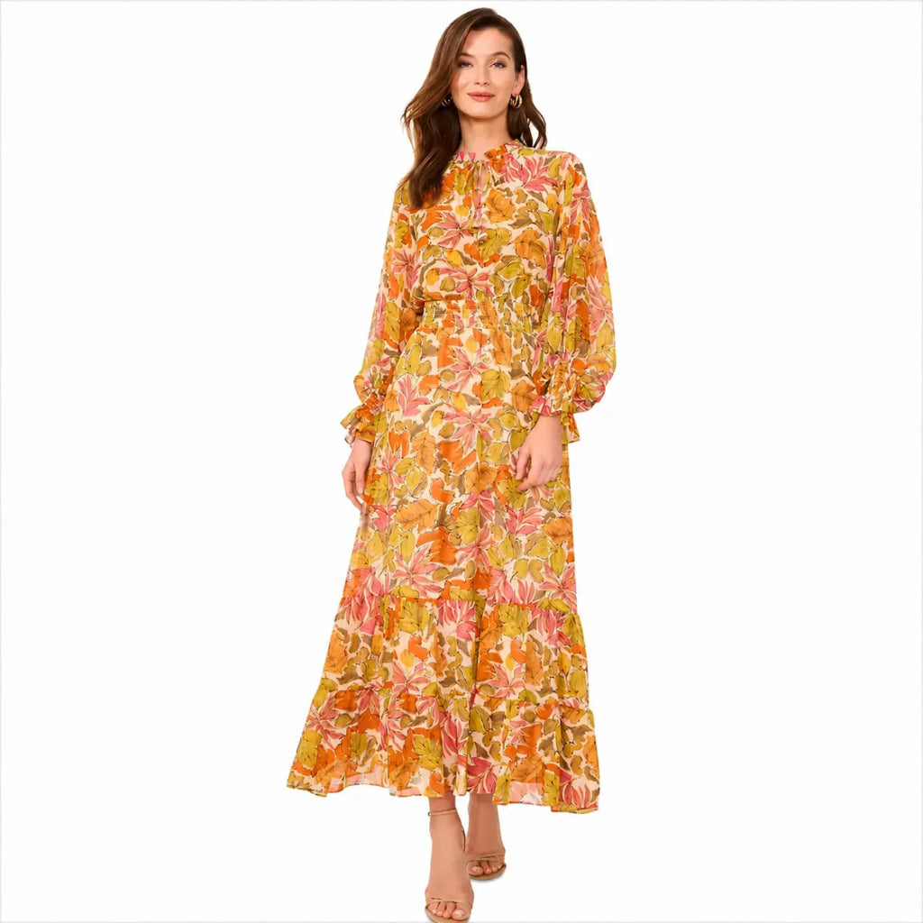 A woman modeling a long-sleeved maxi dress with a vibrant orange, yellow, and pink floral print against a plain white background.