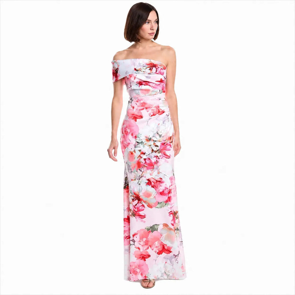 A woman modeling a floor-length off-the-shoulder maxi dress with a pink and red floral print on a white background, set against a plain white studio backdrop.
