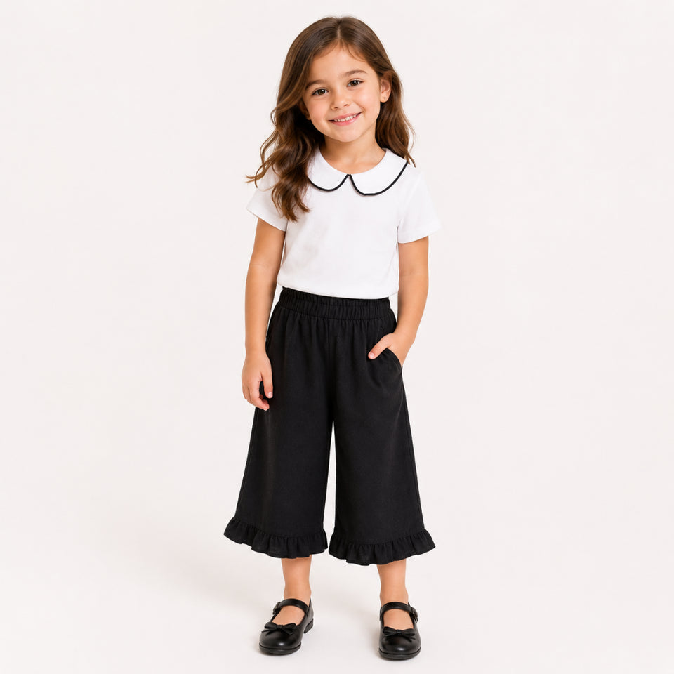 A young girl modeling a white short-sleeve shirt with a black Peter Pan collar and black wide-leg ruffled pants, paired with black Mary Jane shoes, against a plain white studio background.