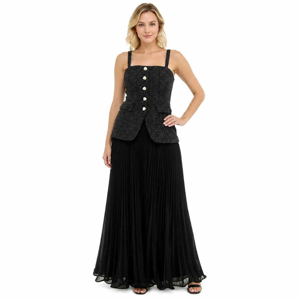 A woman modeling a sleeveless black textured jacket with gold buttons over a long black pleated skirt, shown against a plain white background.
