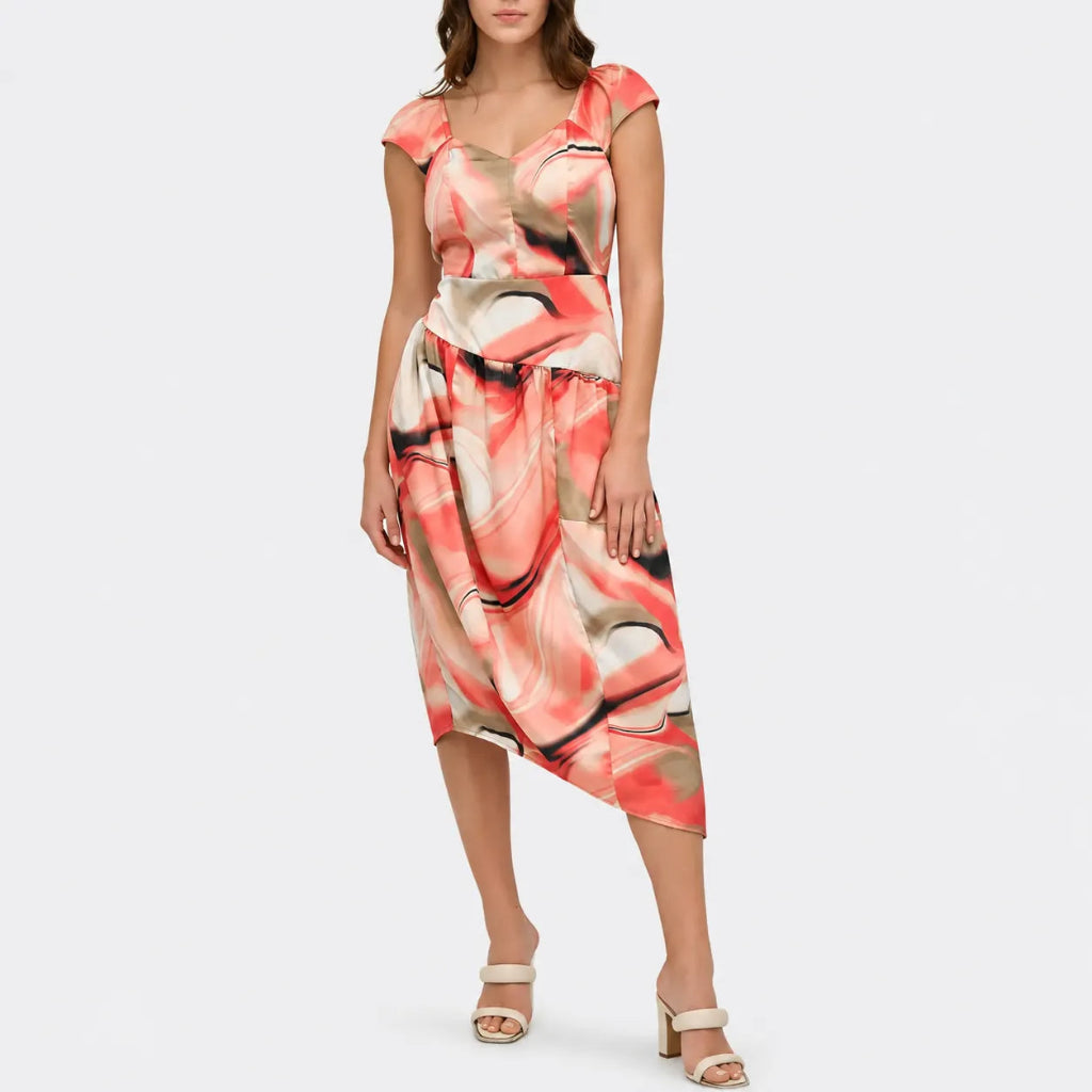 A woman modeling a short-sleeve midi dress with an abstract painterly print in coral pink, beige, and black, paired with beige heeled sandals against a light gray studio background.