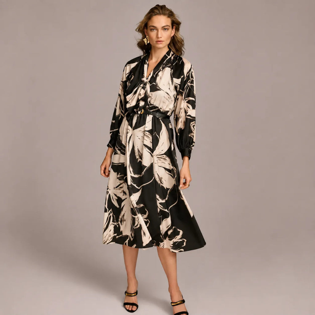 A woman modeling a long-sleeve midi dress with a black and beige abstract floral print, featuring a tie-neck detail and a self-tie belt, set against a solid gray studio background.