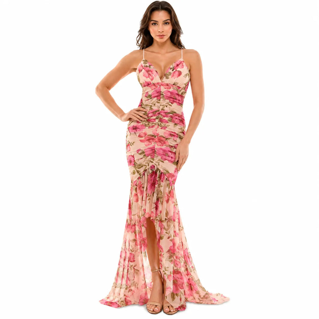 A woman modeling a sleeveless, floor-length floral gown with thin straps, a ruched bodice, and a high slit, shown against a plain white studio background.