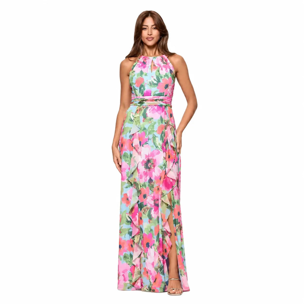 A woman modeling a sleeveless halter-neck maxi dress with a vibrant pink, green, and white floral print, set against a plain white background.