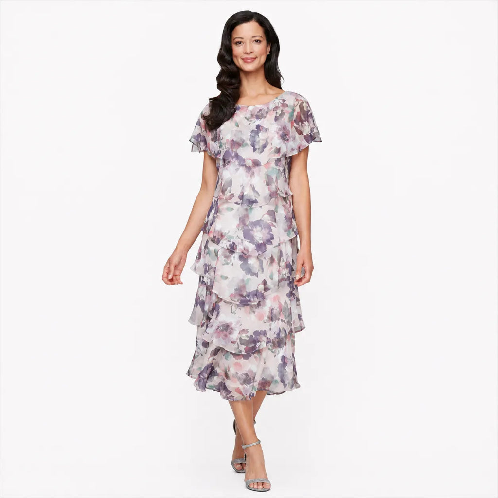 A woman modeling a short-sleeved, tiered midi dress with a watercolor floral print in purple, pink, and green on a white base, set against a plain white background.