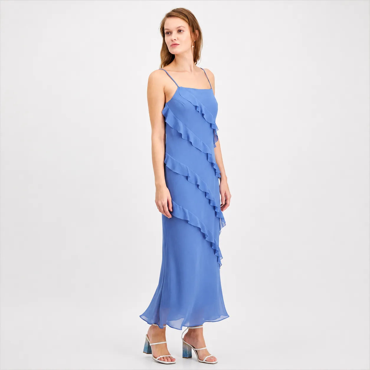 A woman modeling a blue ruffled spaghetti-strap maxi dress against a plain light gray studio background.
