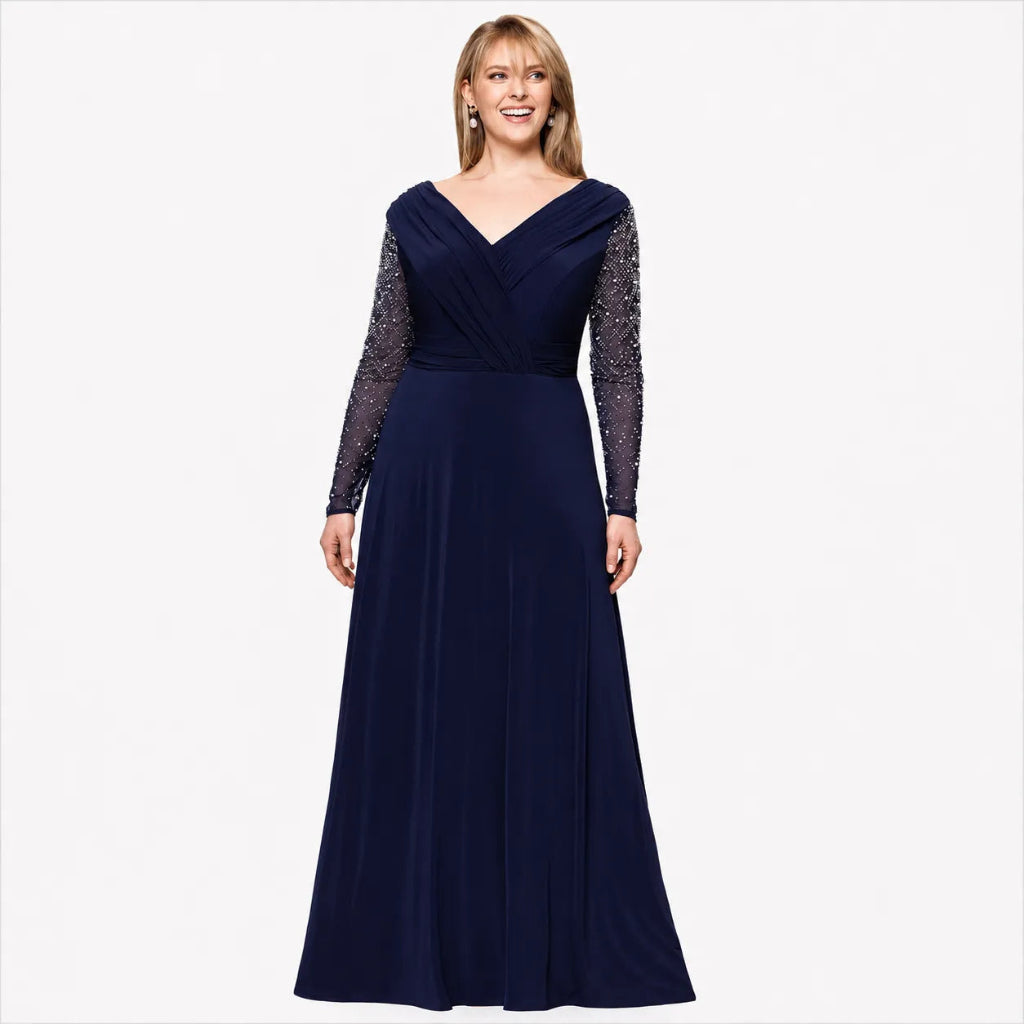 A woman modeling a floor-length navy blue gown with a V-neckline, draped bodice, and long sheer embellished sleeves, photographed against a light gray studio background.