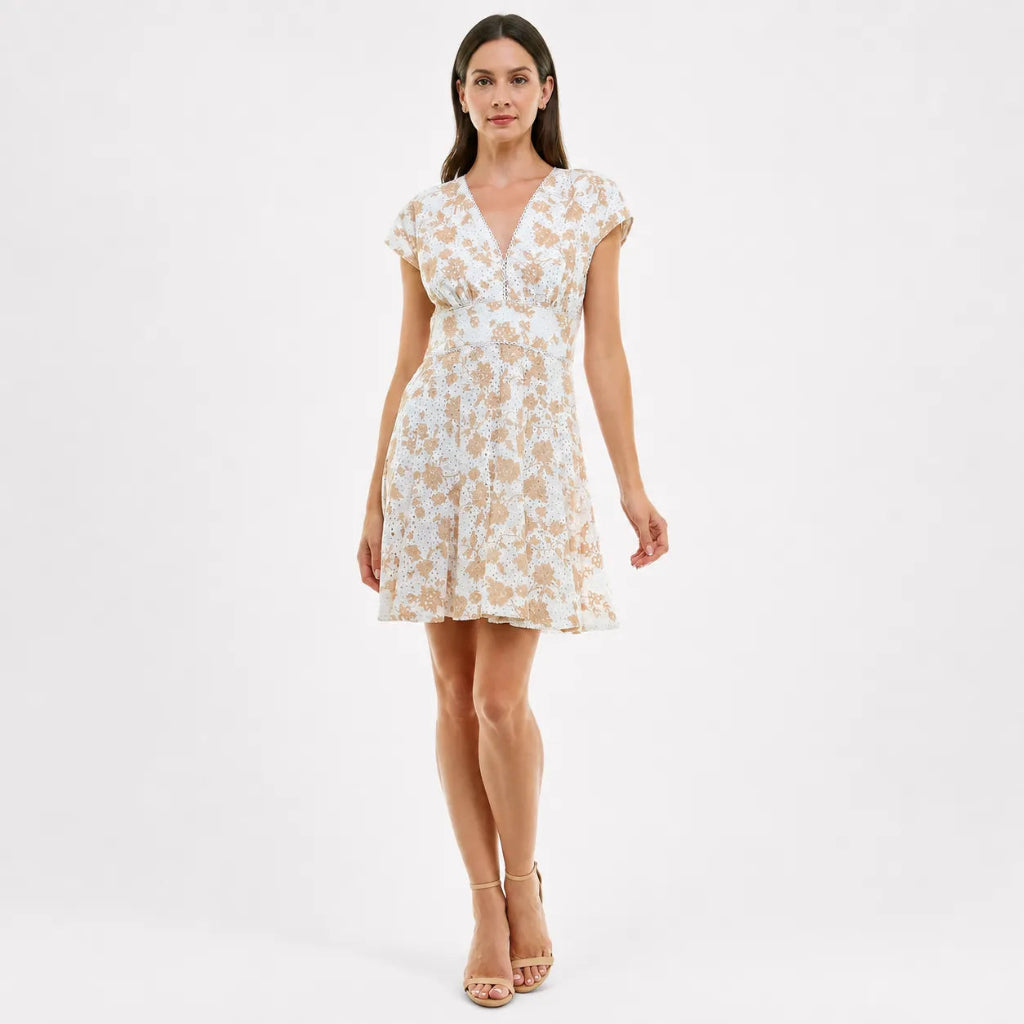 A woman modeling a short white dress with a beige floral pattern, cap sleeves, and a V-neckline against a plain white studio background.