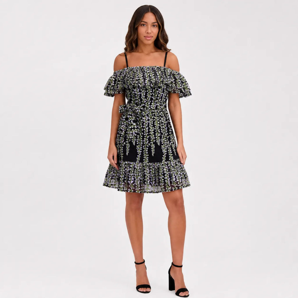 A woman modeling an off-the-shoulder black dress with a dense white floral pattern, featuring ruffled sleeves and a ruffled hem, paired with black high-heeled sandals against a plain white studio background.