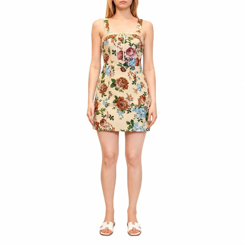 A woman modeling a sleeveless mini dress with a beige background and a colorful floral print, paired with white slide sandals, against a plain white background.