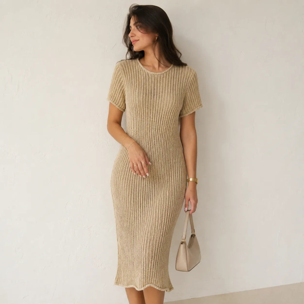 A woman models a beige ribbed short-sleeve midi dress with a scalloped hem, standing against a textured off-white wall and holding a matching beige handbag.